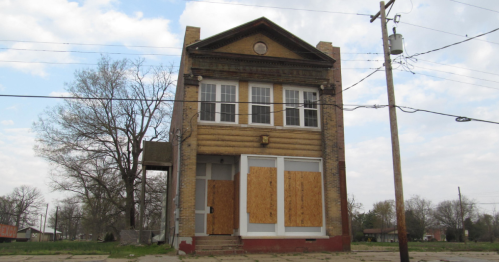 Abandoned two-story building with boarded windows, wooden accents, and a cloudy sky in the background.