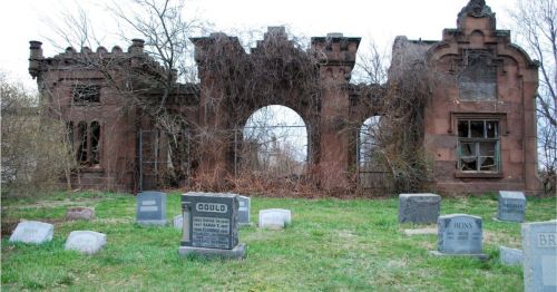 Ruins of a stone building overgrown with vines, surrounded by gravestones in a grassy cemetery.