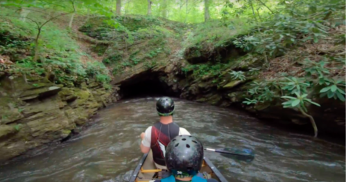 Two people in a canoe navigate a narrow, wooded creek leading to a dark cave entrance. Lush greenery surrounds them.