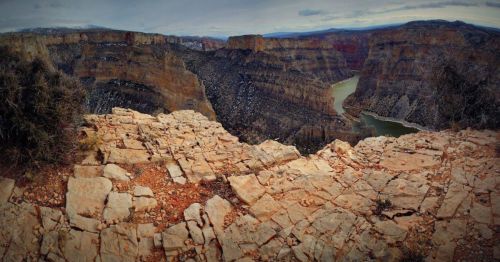 A rocky cliff overlooks a winding river surrounded by steep canyon walls under a cloudy sky.