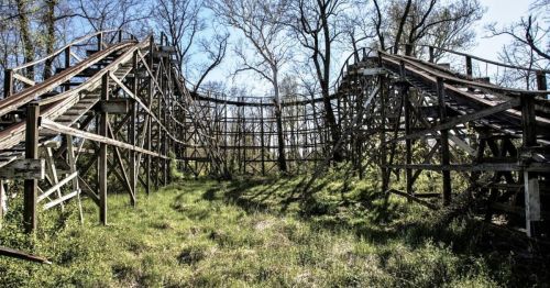 An abandoned wooden roller coaster overgrown with grass and surrounded by trees under a clear blue sky.