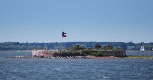 A small island with a flag, surrounded by water, featuring greenery and a distant sailboat on a clear day.