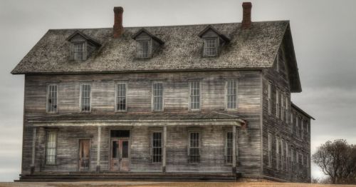A weathered, two-story wooden house with a gray sky overhead, featuring multiple windows and a front porch.