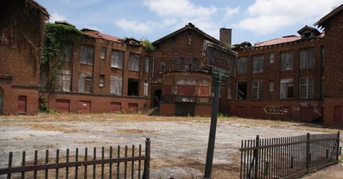 Abandoned brick school building with overgrown vines and a weathered sign, surrounded by a barren lot.