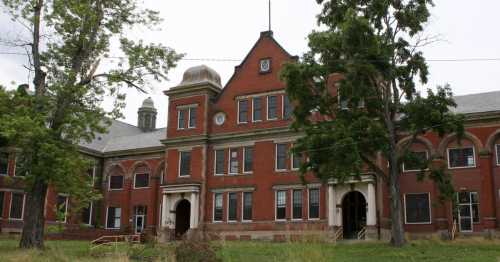 Abandoned red brick building with large windows, surrounded by overgrown grass and trees under a cloudy sky.