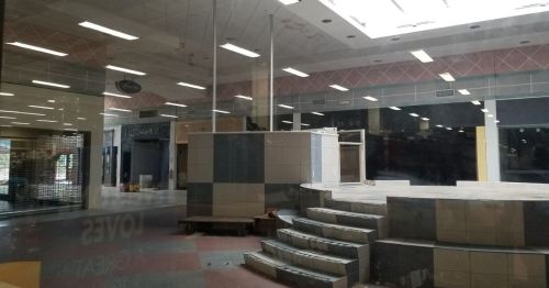 Abandoned retail space with a tiled platform and empty storefronts visible through glass windows.