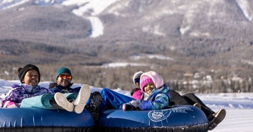 A group of four people enjoying snow tubing on a winter day, with mountains in the background.