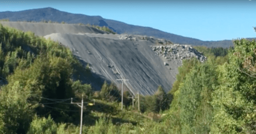 A large gravel slope with trees and power lines in the foreground, set against a mountainous backdrop under clear blue skies.
