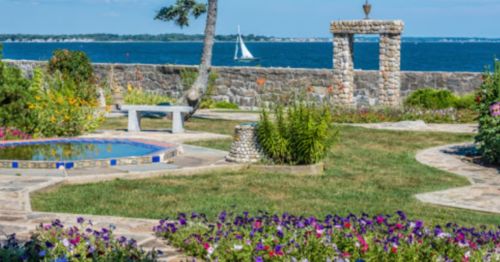 A scenic garden with colorful flowers, stone pathways, and a view of the ocean with a sailboat in the distance.