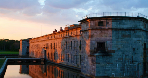 Historic stone building with a moat, reflecting sunset colors, surrounded by green landscape and cloudy sky.