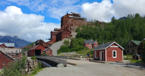 Historic mining buildings in a mountainous landscape, with a blue sky and scattered clouds above.