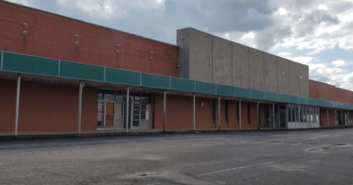Abandoned building with red brick and gray concrete, featuring a green awning and empty storefronts under a cloudy sky.