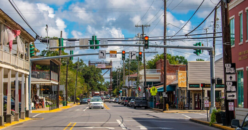 A small town street with shops, traffic lights, and a blue sky with clouds. Cars are parked along the road.