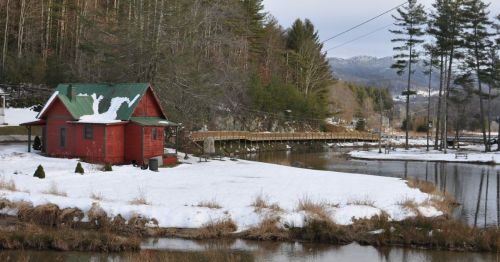 A red cabin with a green roof sits by a snowy river, surrounded by trees and mountains in the background.
