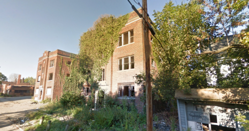 Abandoned brick building overgrown with vegetation, surrounded by a neglected lot and power lines.