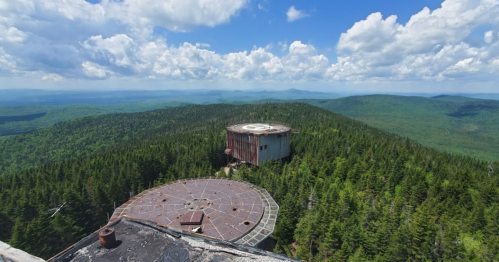Aerial view of two large, circular structures atop a green mountain, surrounded by dense forest and blue sky with clouds.