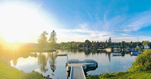 A serene lakeside scene at sunset, featuring a dock, boats, and trees reflecting on calm water.