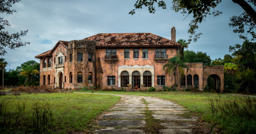 Abandoned mansion with a weathered facade, surrounded by overgrown grass and trees under a cloudy sky.