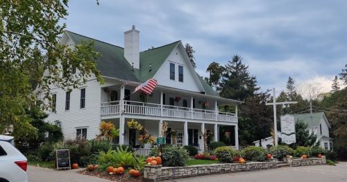 A charming white house with a green roof, decorated with pumpkins and an American flag, surrounded by lush greenery.