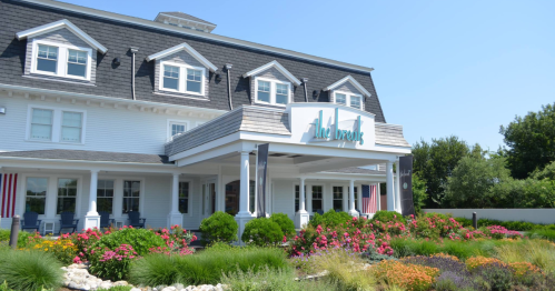 A charming hotel with a white facade, blue signage, and colorful flower beds under a clear blue sky.