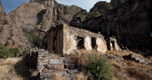 Ruins of a stone building surrounded by dry grass and steep rocky cliffs under a clear sky.