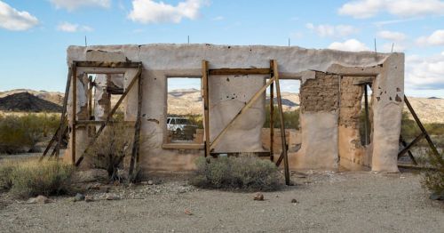 Abandoned, partially collapsed building in a desert landscape, surrounded by sparse vegetation and mountains in the background.