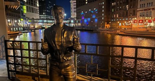 A bronze statue of a smiling man giving a thumbs-up, with a river and city lights in the background at night.