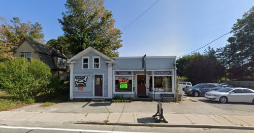 A small blue storefront with signs, surrounded by trees and parked cars on a sunny day.