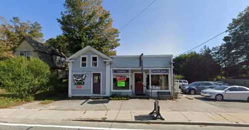 A small blue storefront with signs, surrounded by trees and parked cars on a sunny day.