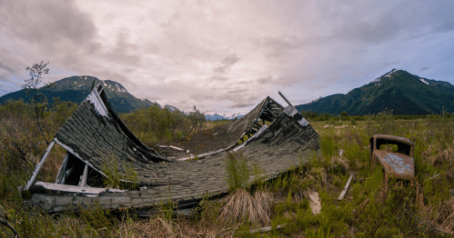 A dilapidated building lies in a grassy field, surrounded by mountains under a cloudy sky. An old rusted vehicle is nearby.