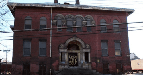 Abandoned red brick building with large windows and a sign reading "KNOW THY SELF" on the front entrance.