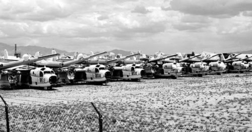 A row of military helicopters parked in a desert landscape, with mountains and cloudy skies in the background.