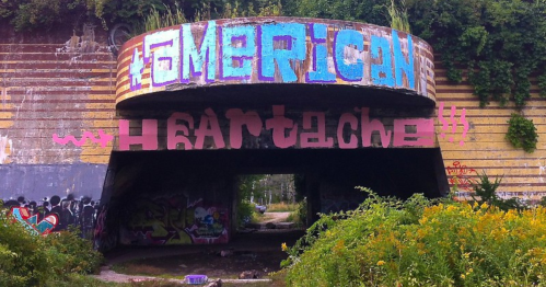 Graffiti-covered concrete structure with the words "American Heartache" and vibrant colors, surrounded by greenery.