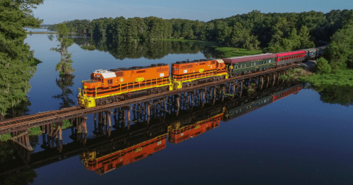 A train crosses a wooden trestle over a calm river, surrounded by lush greenery and reflecting in the water.