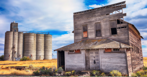An abandoned wooden house beside tall, cylindrical grain silos under a cloudy sky, surrounded by dry grass.