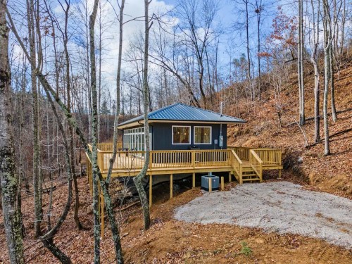A small cabin with a deck, surrounded by trees and autumn leaves, set on a hillside under a blue sky.