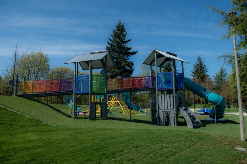 Colorful playground structure with slides and climbing areas, surrounded by green grass and trees under a blue sky.