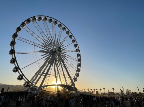 A silhouette of a Ferris wheel against a sunset sky, with people gathered below and palm trees in the background.