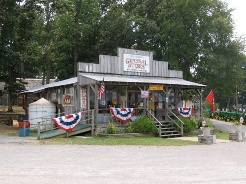 A rustic general store with a wooden facade, flags, and bunting, surrounded by trees and a gravel path.