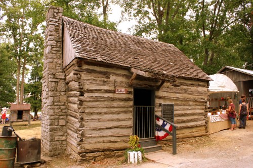 A rustic log cabin with a stone chimney, surrounded by trees and a small market area.