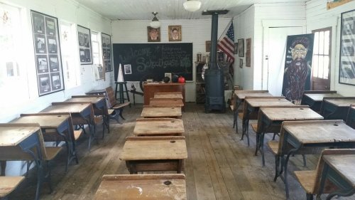 A vintage classroom with wooden desks, a chalkboard, and historical photos on the walls. An American flag is displayed.