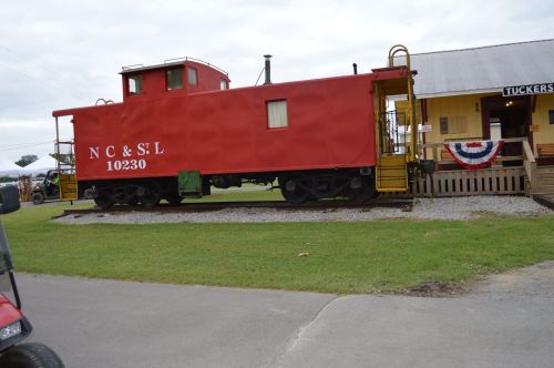 A red caboose labeled "N C & S L 10230" parked beside a yellow building with patriotic decorations.