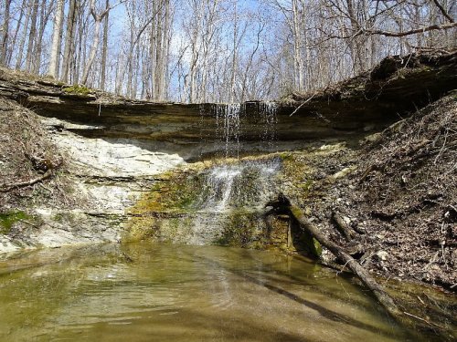 A serene waterfall cascading over rocky terrain into a calm pool, surrounded by bare trees and blue sky.
