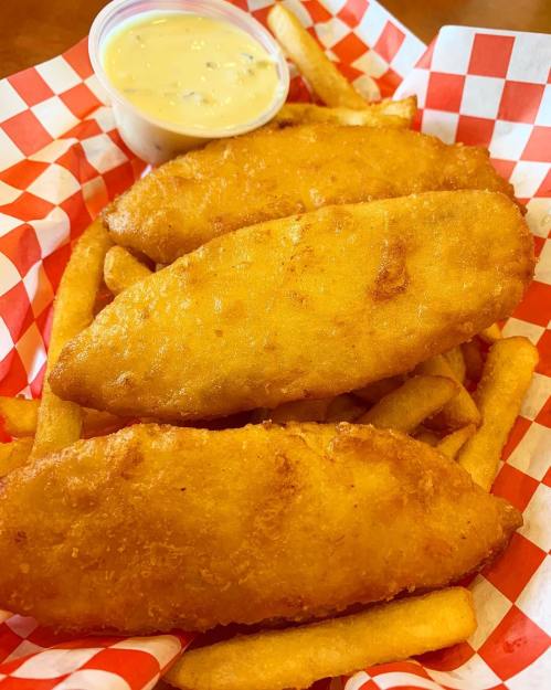 Three golden-brown fried fish fillets served with French fries and a small cup of dipping sauce in a red-checkered basket.