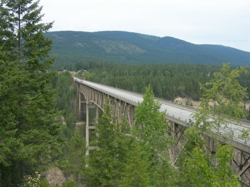 A long bridge spans a valley, surrounded by dense green forests and mountains under a cloudy sky.