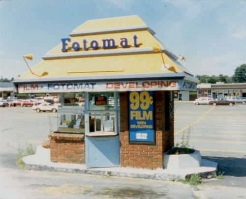 A small, vintage Fotomat kiosk in a parking lot, advertising 99¢ film developing services.