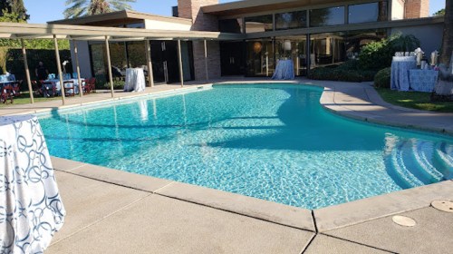 A clear blue pool surrounded by a patio and tables, set in a sunny outdoor space with modern architecture.