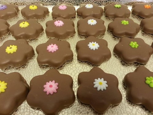 A tray of chocolate-covered flower-shaped cookies, decorated with colorful icing flowers in pink, white, and green.