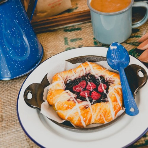 A blueberry pastry in a cast iron dish, with a blue spoon, coffee mug, and a blue kettle in the background.
