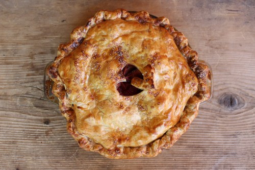A golden-brown pie with a flaky crust, featuring a decorative top and a small vent hole, resting on a wooden surface.
